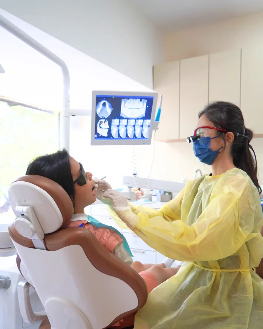 Dentist in a surgical gown and loupes at the chair with the X-ray monitor above